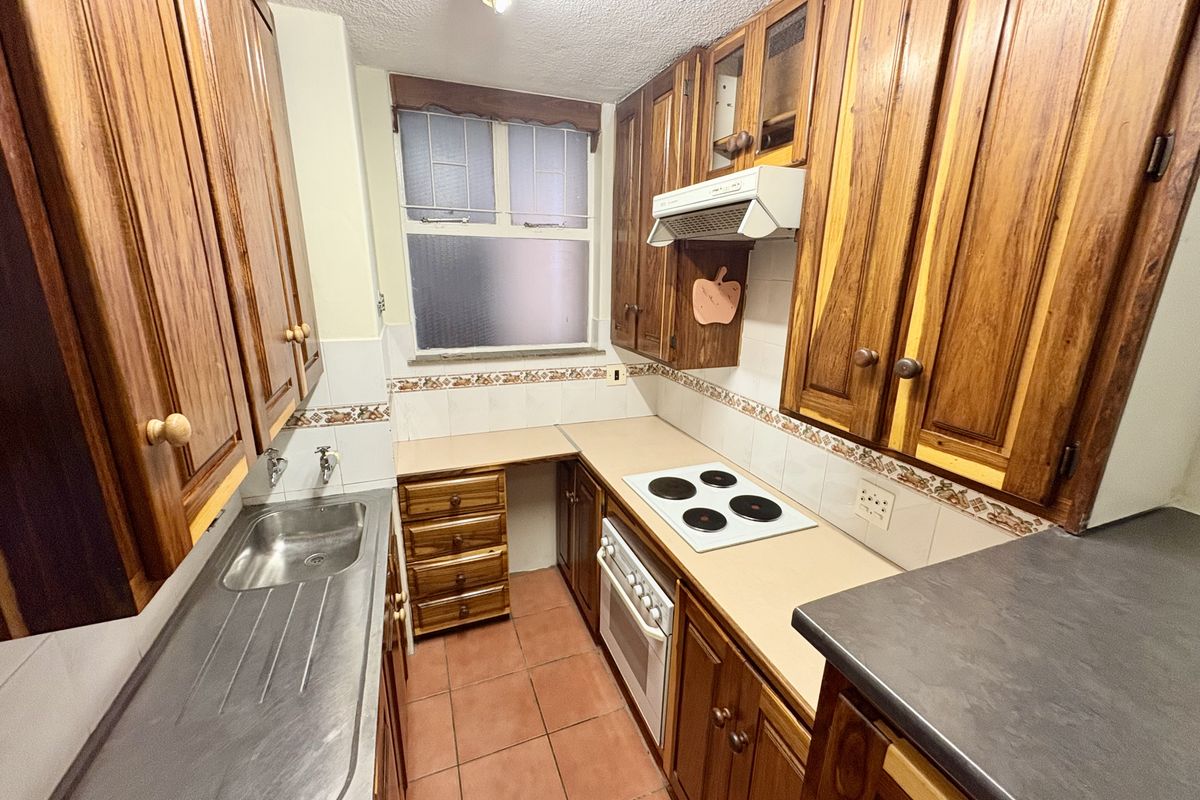 A compact kitchen featuring traditional wooden cabinetry, a built-in oven with electric hob, stainless steel sink, and tiled flooring.