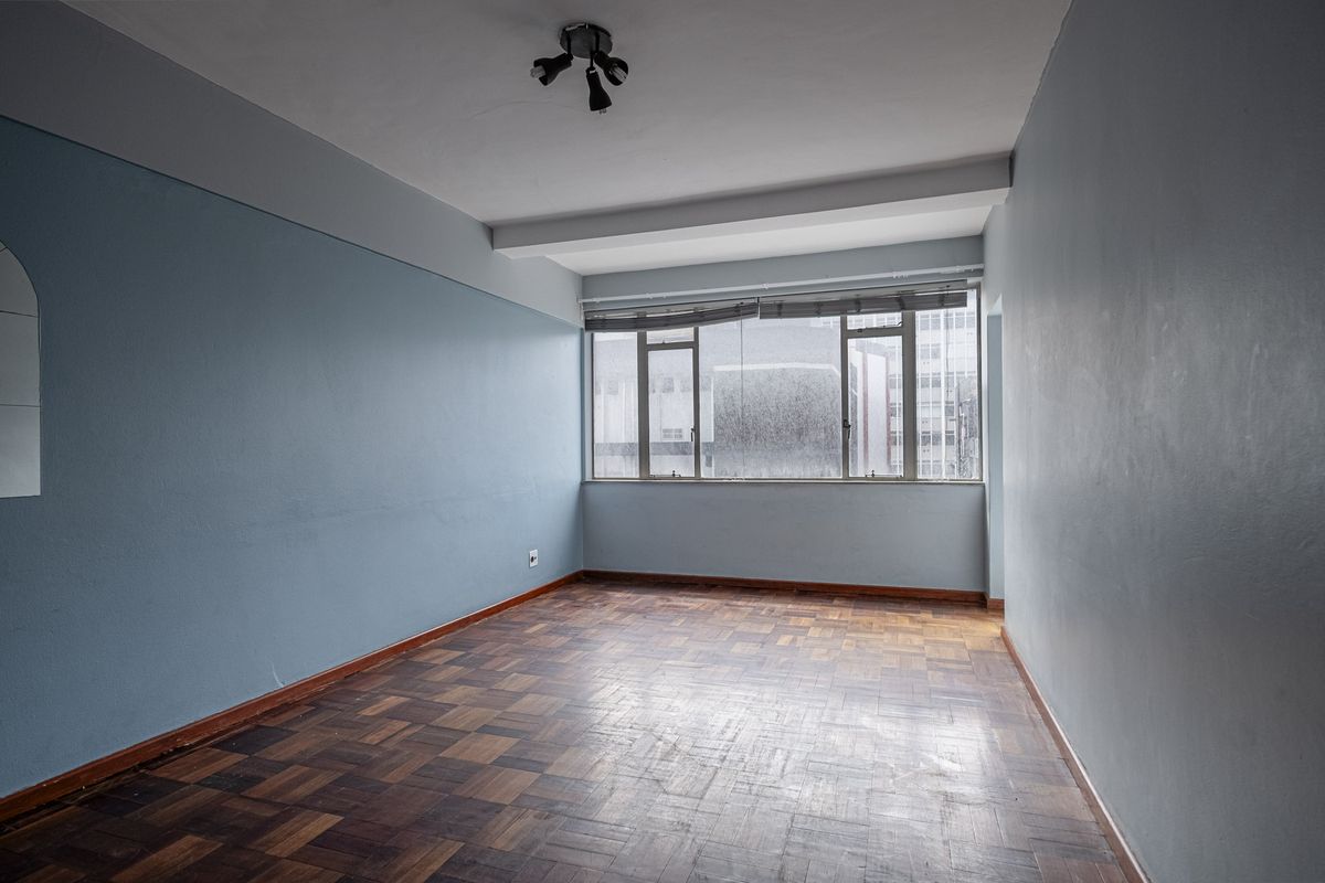 An empty living room featuring original parquet flooring, light blue walls, and large windows providing natural light.