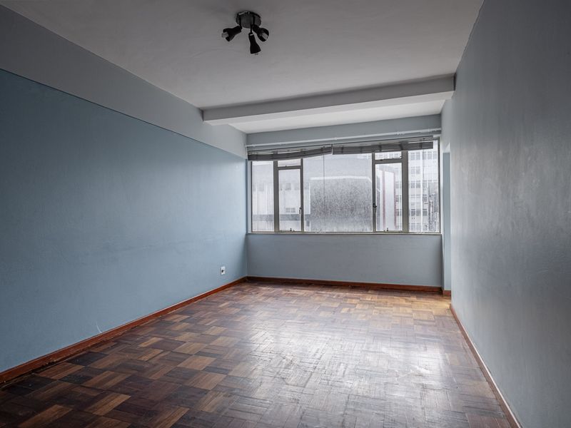 An empty living room featuring original parquet flooring, light blue walls, and large windows providing natural light.