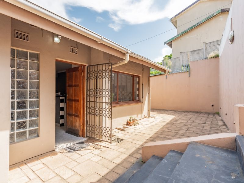 A paved rear patio area featuring a security gate, glass block window, and concrete stairs leading to the exterior entrance.