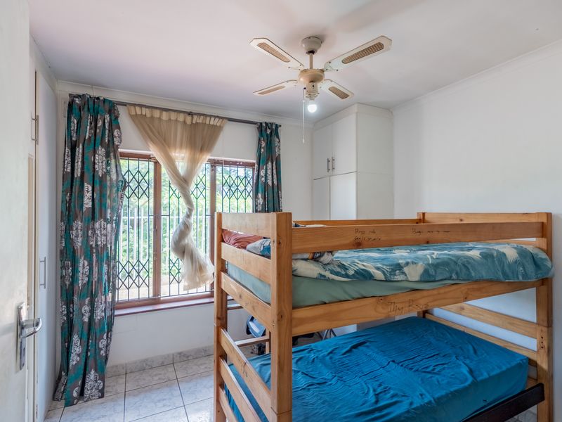 A functional bedroom featuring tiled flooring, built-in cupboards, and a ceiling fan, with a window protected by a security gate.