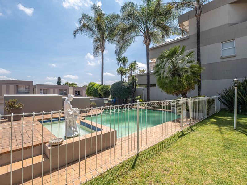 A well-maintained communal swimming pool area featuring a paved surround, decorative water feature, and established garden landscaping within the apartment complex.