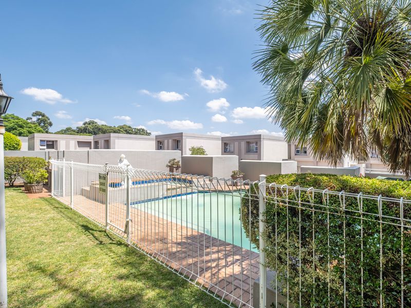 A well-maintained communal swimming pool area featuring paved surrounds, metal security fencing, and established garden landscaping within an apartment complex.