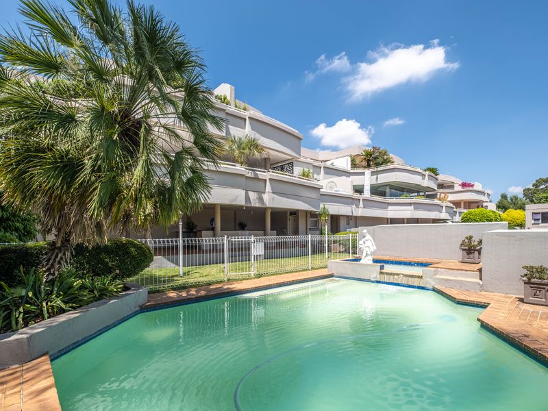 A communal swimming pool area featuring a water feature, brick coping, and white fencing, set against the backdrop of a multi-level apartment building.