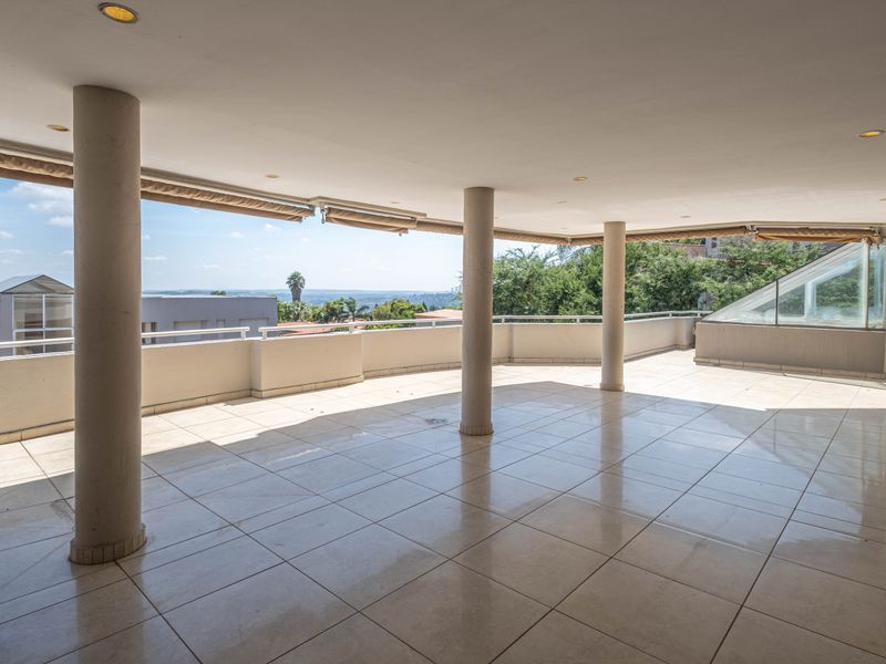 A spacious, covered balcony featuring tiled flooring, structural columns, and retractable awnings, offering an expansive outdoor area with scenic views.