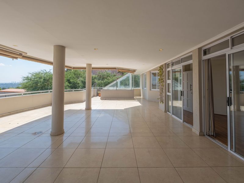 A spacious, covered balcony featuring tiled flooring, structural columns, and large sliding glass doors, offering an expansive outdoor living area with scenic views.