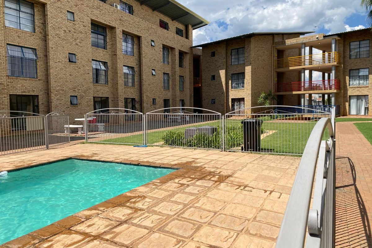 Communal pool area featuring a paved deck, metal safety fencing, and landscaped garden space set against a multi-story brick apartment complex.