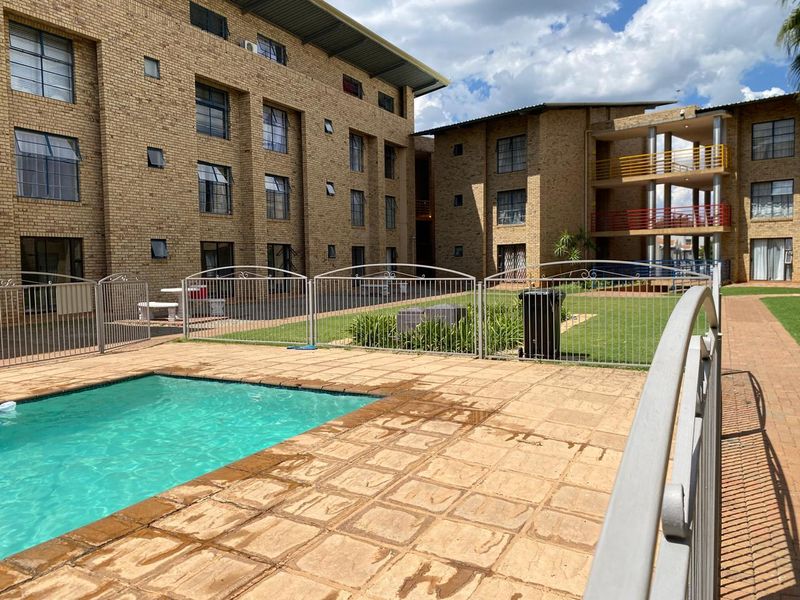 Communal pool area featuring a paved deck, metal safety fencing, and landscaped garden space set against a multi-story brick apartment complex.