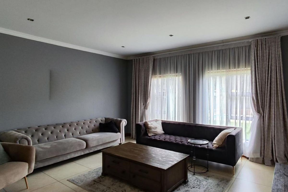 A spacious living room featuring large windows with floor-to-ceiling curtains, recessed downlighting, and neutral tiled flooring.