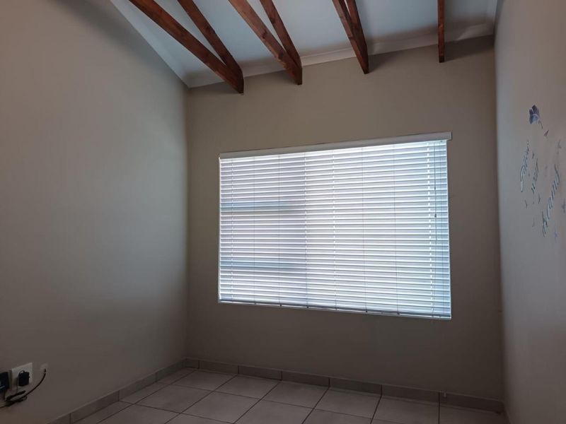 A bright, empty bedroom featuring exposed timber ceiling beams, neutral wall tones, tiled flooring, and fitted venetian blinds.