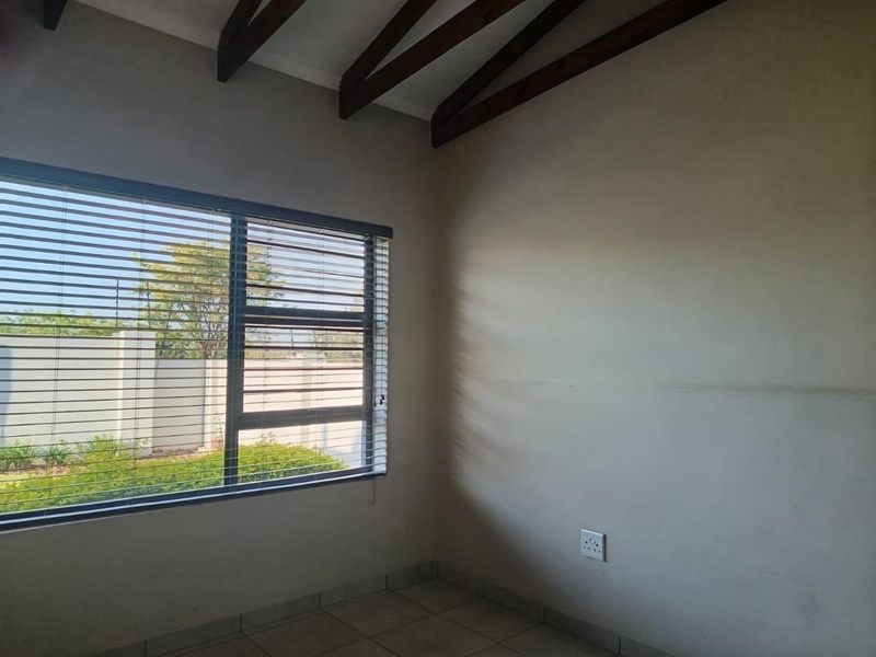 A vacant bedroom featuring exposed timber ceiling beams, tiled flooring, and a large window with fitted venetian blinds overlooking the garden.
