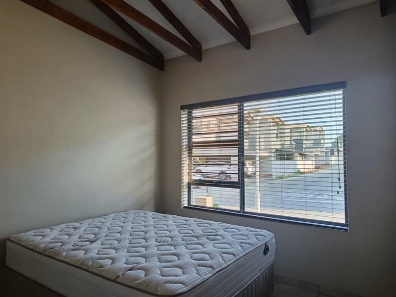A bedroom featuring exposed timber ceiling beams, tiled flooring, and large windows fitted with venetian blinds, offering a view of the complex.
