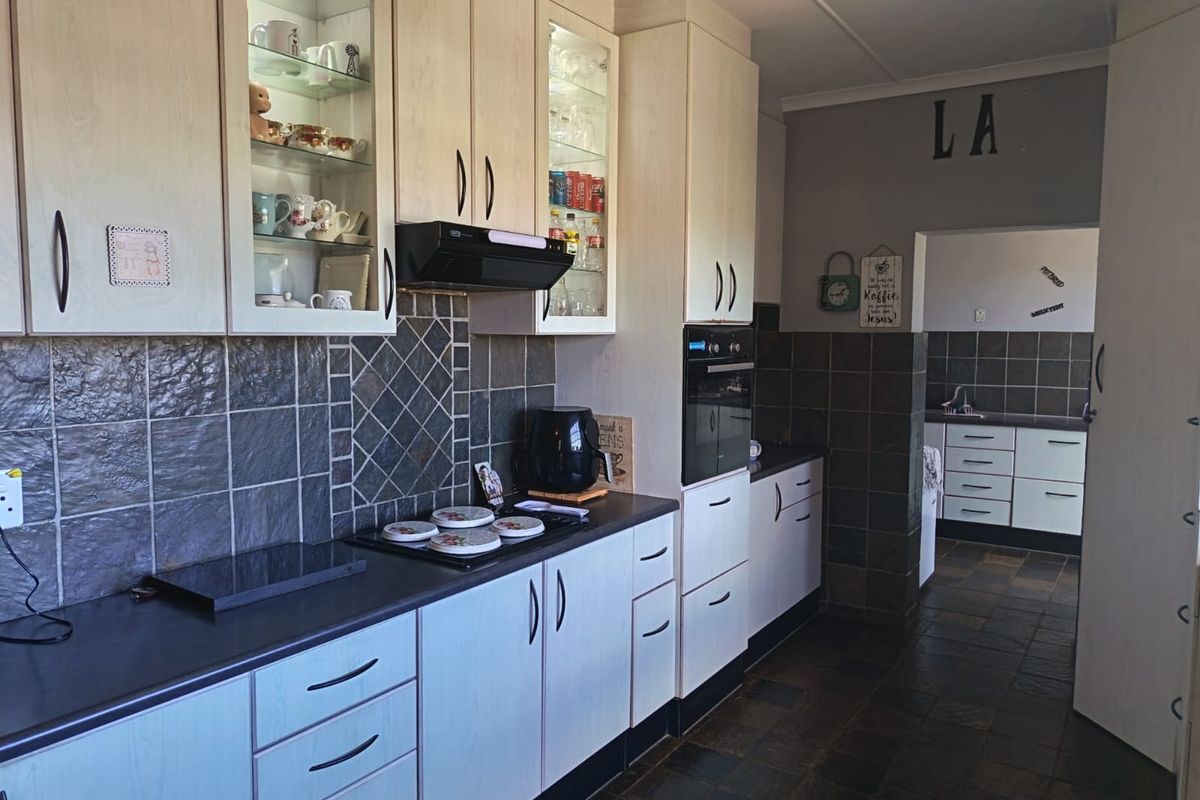 A functional kitchen featuring light-toned cabinetry, dark tiled splashbacks, built-in oven, and hob, with an adjacent scullery area visible in the background.