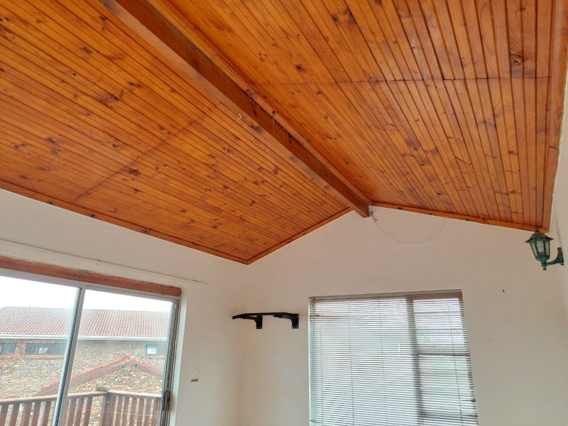 Living area featuring a vaulted timber tongue and groove ceiling with exposed beam, sliding glass doors, and large windows with venetian blinds.