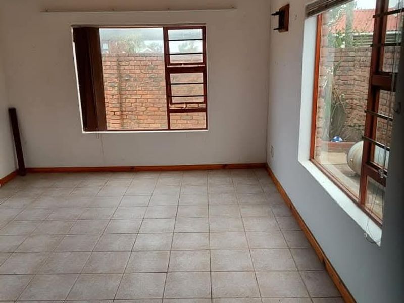 An empty living room featuring exposed timber ceiling beams, tiled flooring, and large windows with security bars overlooking the rear garden area.