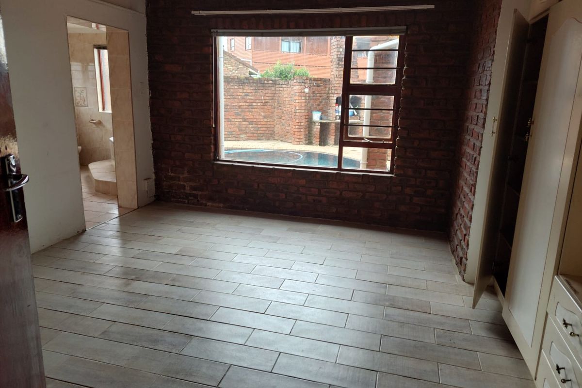 An empty bedroom featuring exposed brick walls, modern tiled flooring, and built-in cupboards, with a view overlooking the pool area.