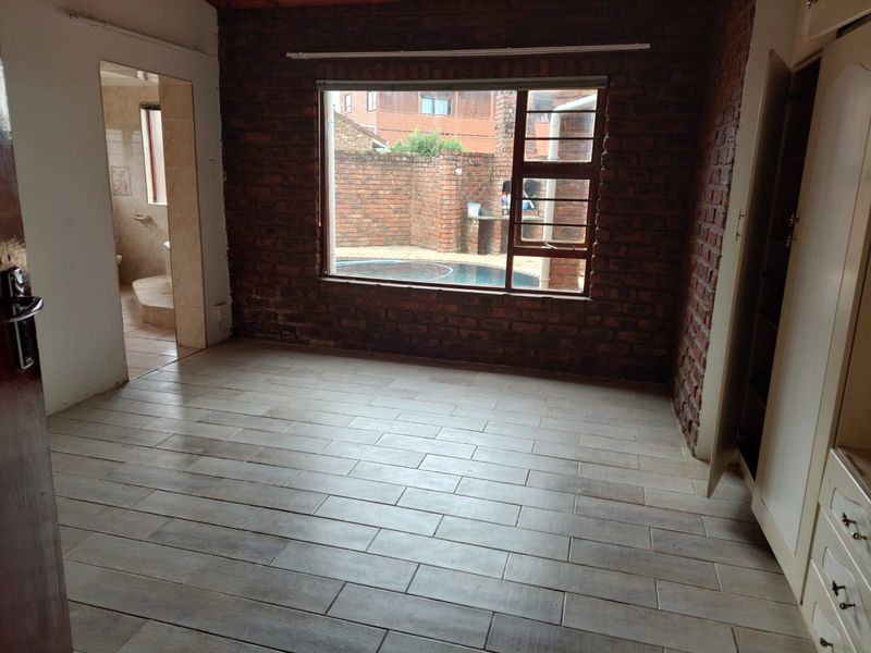 An empty bedroom featuring exposed brick walls, modern tiled flooring, and built-in cupboards, with a view overlooking the pool area.