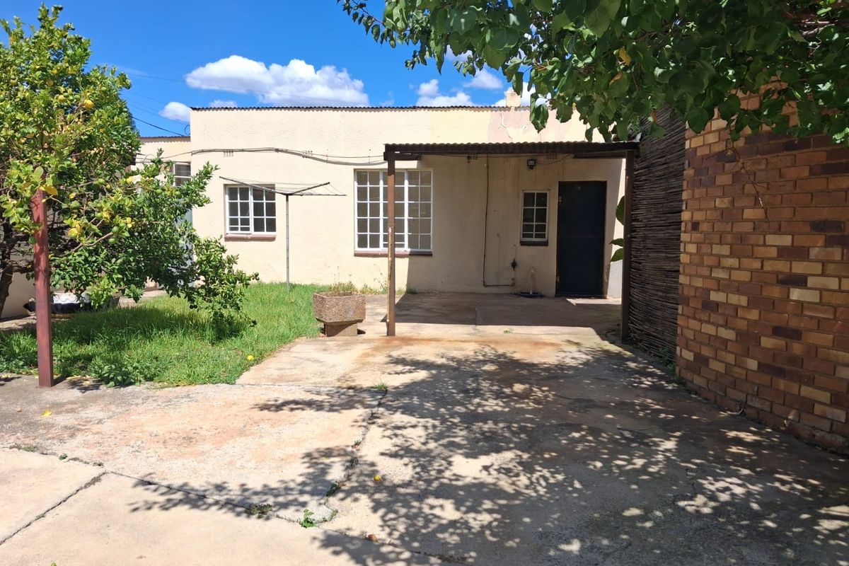 Rear exterior view of the property featuring a concrete patio area, covered veranda, and established garden space with brick and plastered wall finishes.
