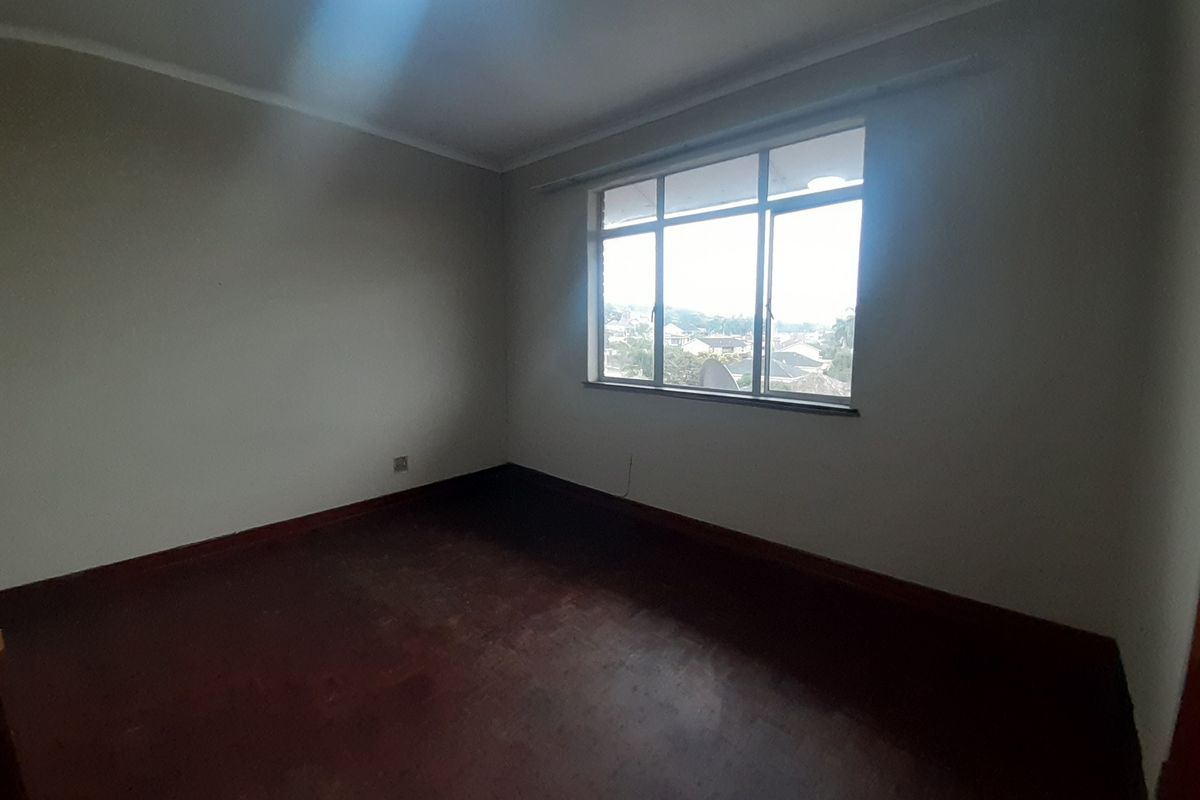 An empty bedroom featuring dark wooden flooring, neutral walls, and a large window providing natural light and an exterior view.