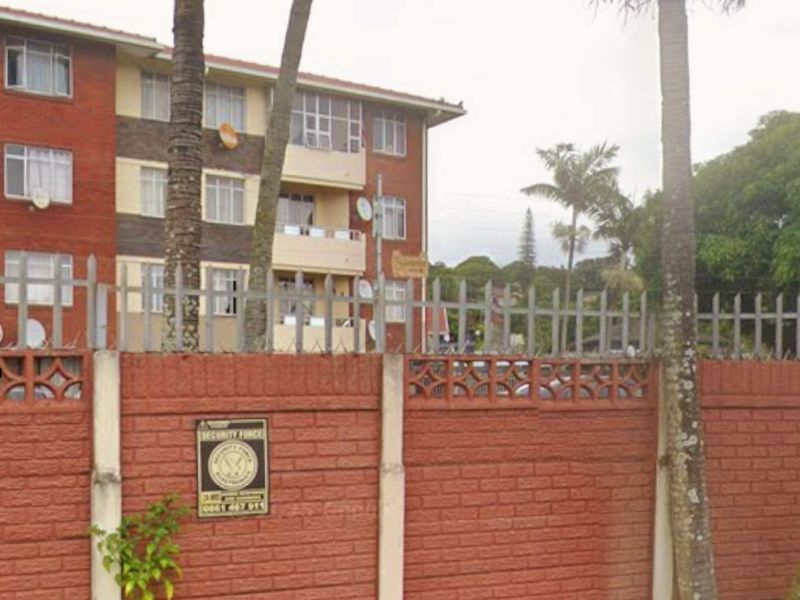Front view of a multi-story brick apartment building featuring a secure perimeter wall with steel palisade fencing and established palm trees.