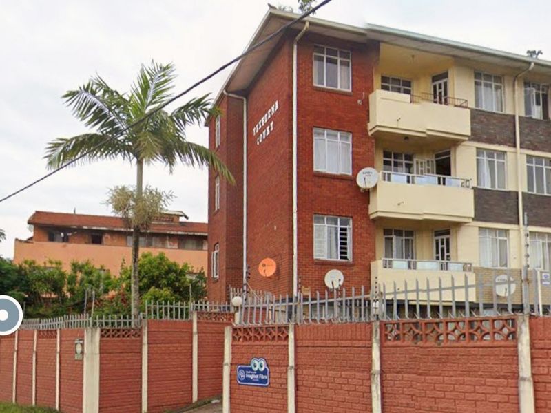 Front exterior view of a multi-story brick apartment building featuring balconies, secure perimeter walling, and electric fencing.
