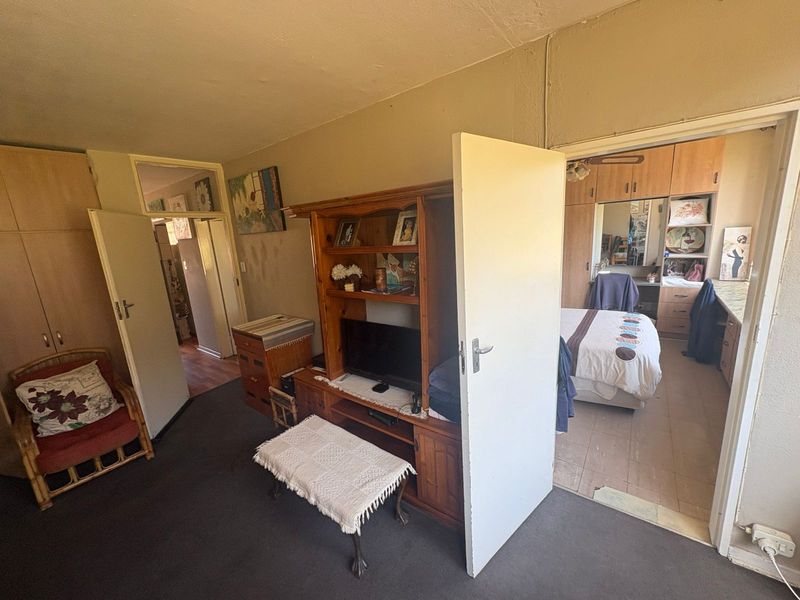 A bedroom featuring built-in cupboards and tiled flooring, with an adjacent room visible through the doorway. The space requires cosmetic updates and maintenance.
