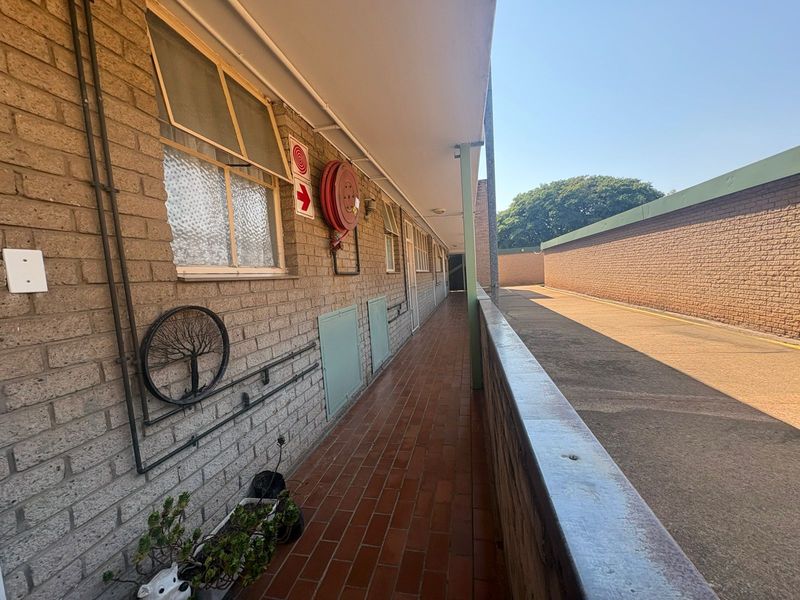 An exterior open-air walkway featuring brick walls, tiled flooring, and a fire safety hose reel, providing access to the flat units.