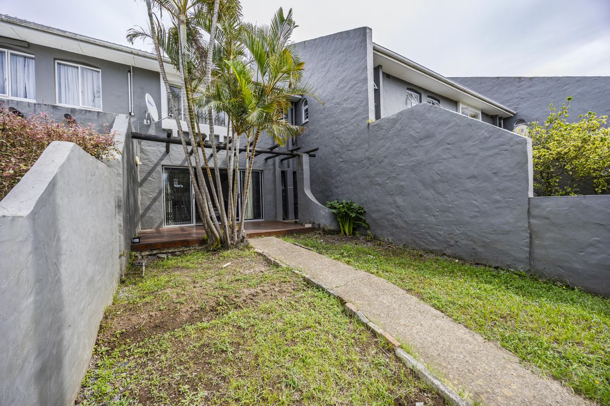 Rear view of a townhouse featuring a small garden, a concrete pathway, and a wooden deck area leading to sliding glass doors.