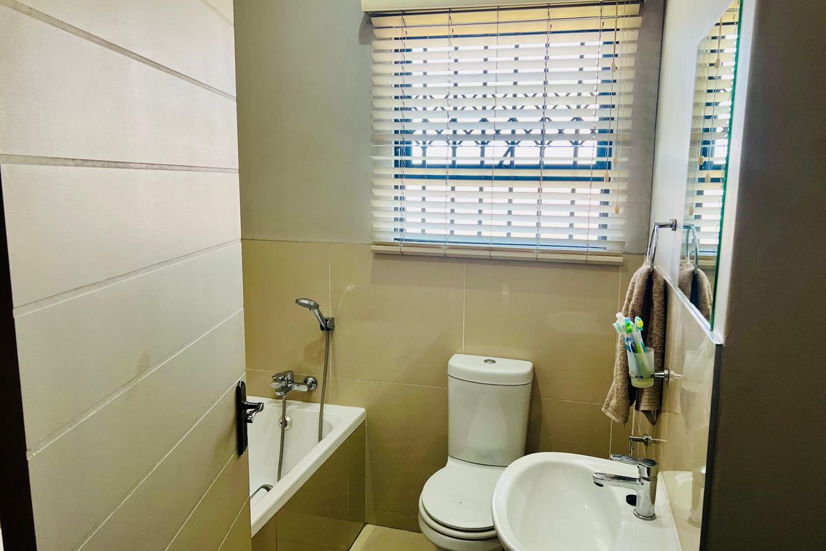 A functional bathroom featuring a bathtub with handheld shower, pedestal sink, toilet, and neutral wall tiling with venetian blinds for privacy.