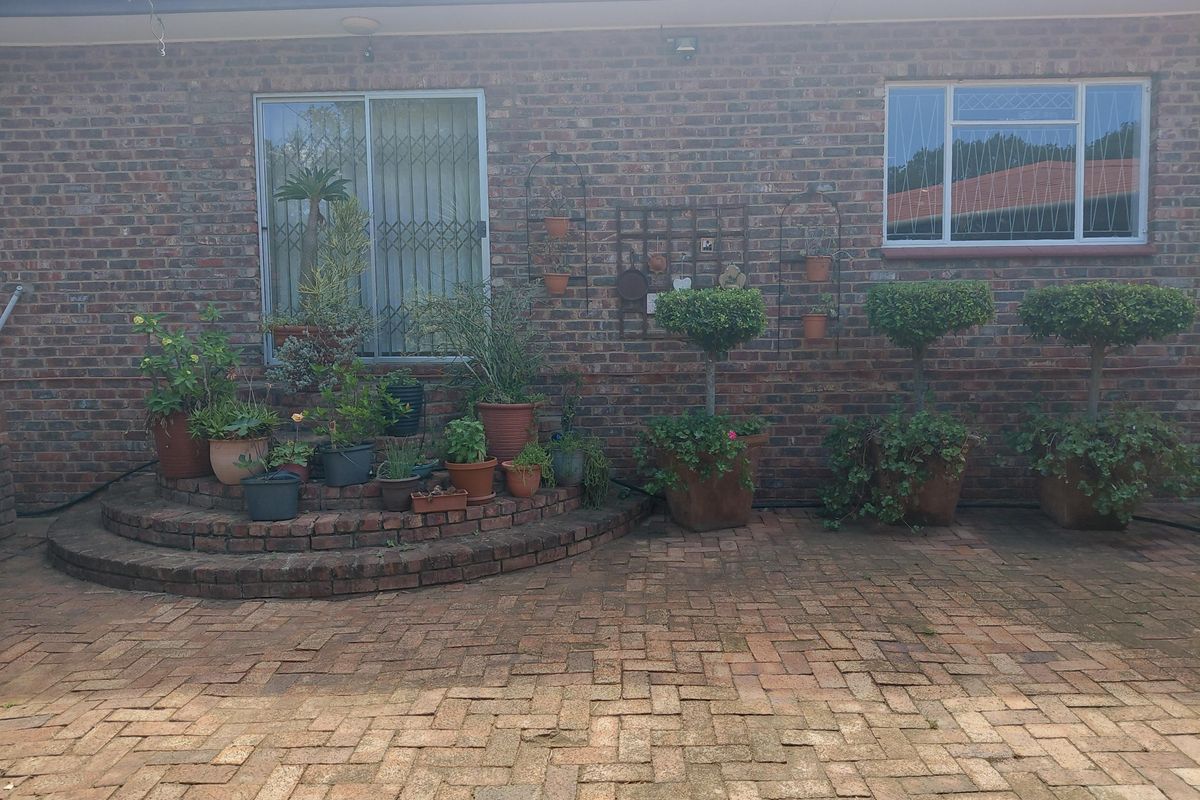 A rear exterior view featuring a brick facade, a paved patio area, and a tiered brick planter with established greenery.