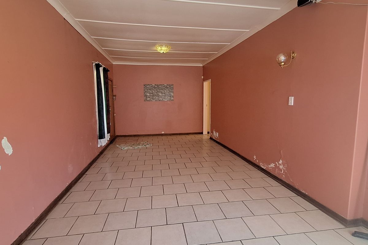 An empty living room featuring tiled flooring, exposed beam ceiling, and wall-mounted lighting fixtures, requiring cosmetic updates to the wall finishes.