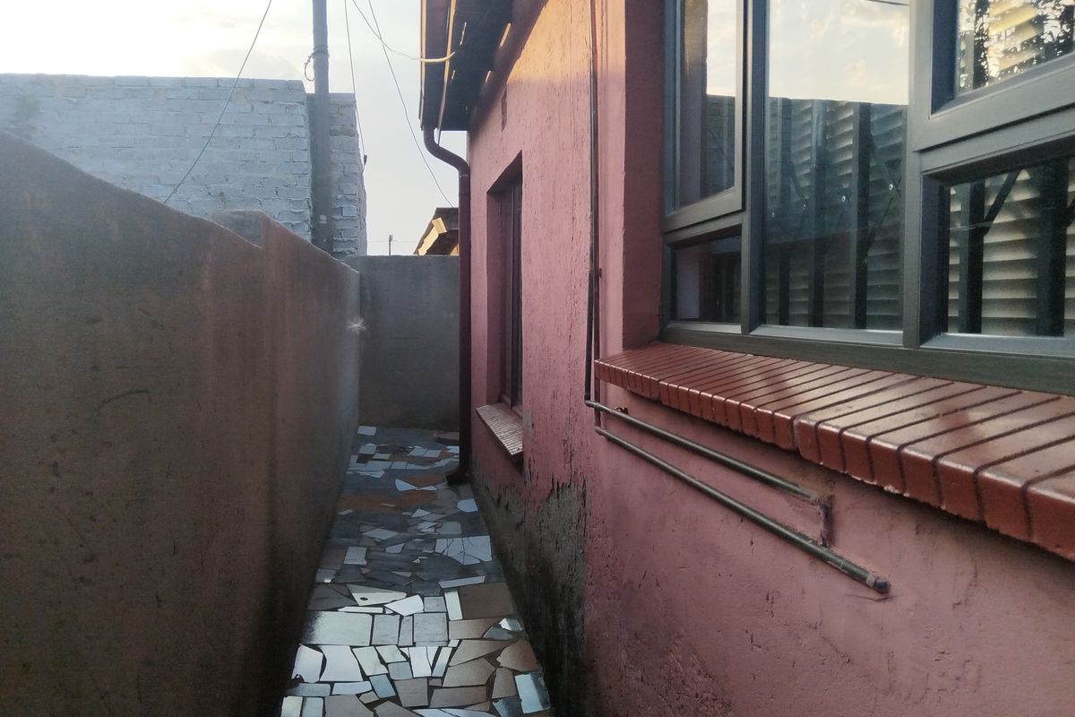 A narrow side passage featuring a tiled walkway, aluminium windows with brick sills, and a boundary wall, showing signs of exterior plaster deterioration.