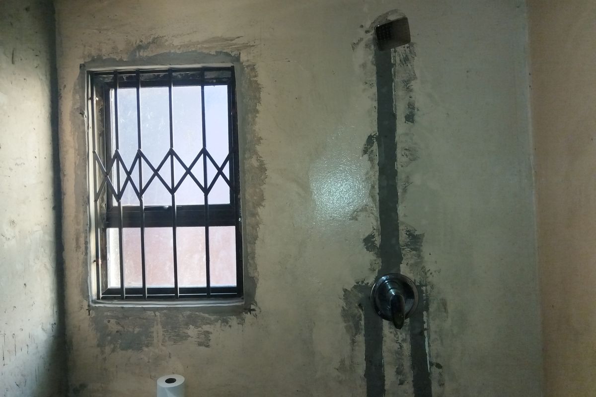 A bathroom in a state of renovation with exposed plumbing channels and unfinished wall plastering, featuring a shower fixture and a window with security gate.