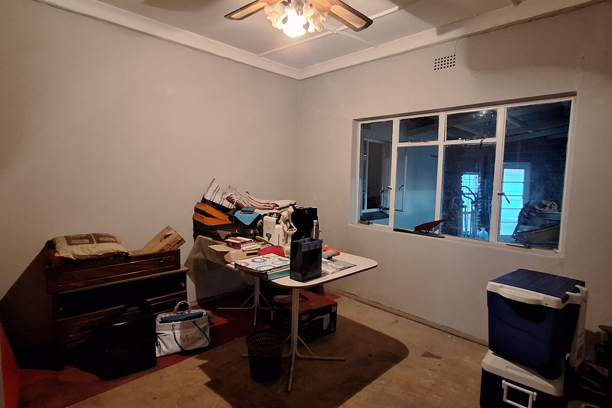 A bedroom space featuring a ceiling fan and large window, currently used as a storage area with worn flooring and dated finishes.