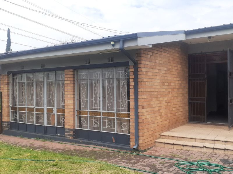Front elevation of a brick house featuring metal security gates, tiled entrance steps, and large metal-framed windows overlooking a paved area and lawn.
