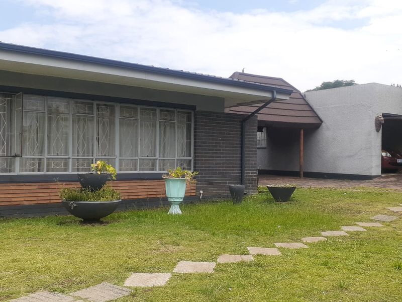 Front view of a single-story house featuring a brick facade, large windows with security bars, a paved driveway, and a carport.