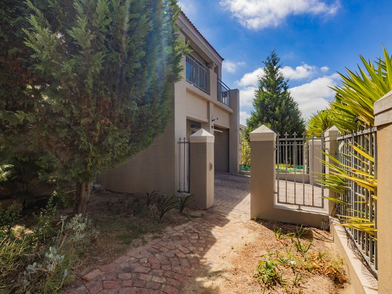 Front exterior view of the property featuring a brick paved driveway, secure metal gate, masonry boundary wall, and an upper-level balcony.