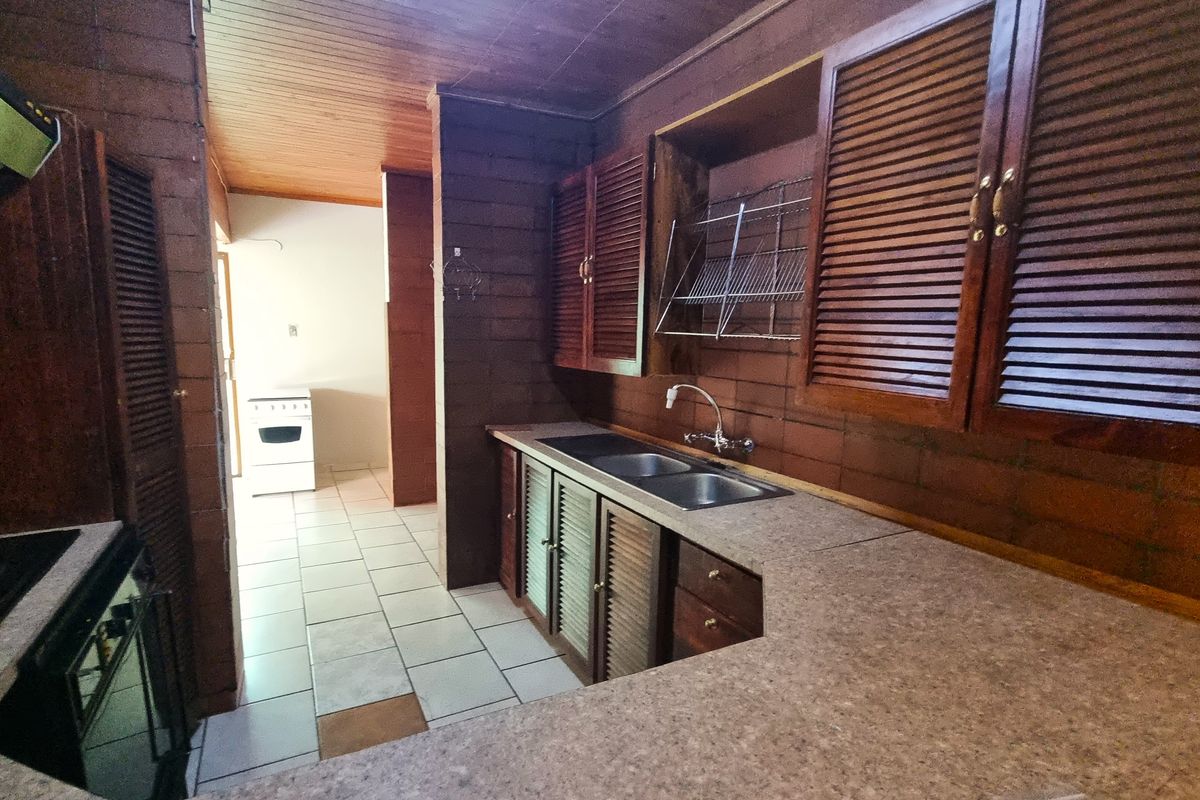 A dated kitchen featuring dark timber louvered cabinetry, a double basin sink, and tiled flooring, offering a functional layout with potential for modernization.