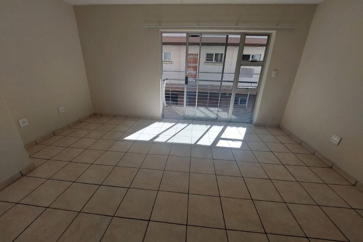 An empty, sunlit living area featuring neutral tiled flooring and a sliding glass door providing access to an outdoor balcony.