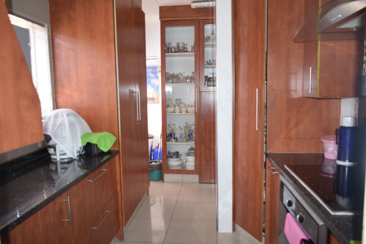 A functional kitchen featuring warm-toned wood cabinetry, granite countertops, integrated appliances, and a glass-front display cabinet, finished with neutral floor tiles.