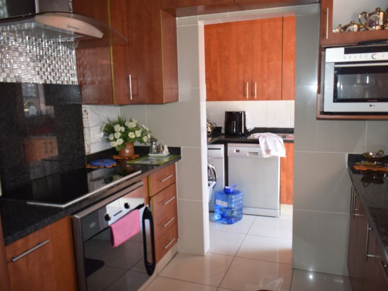 A functional kitchen featuring wood-finish cabinetry, granite countertops, integrated appliances, and a tiled backsplash, with an adjoining scullery area.