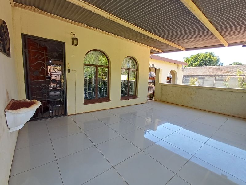 A covered patio area featuring tiled flooring, arched windows with security bars, and a corrugated metal roof, providing a functional outdoor space.