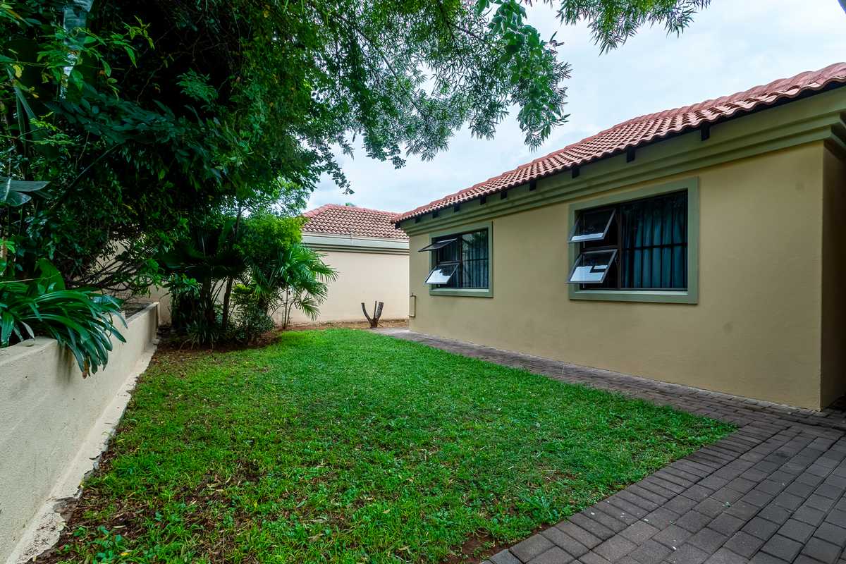 A private rear garden featuring a paved walkway, established greenery, and a well-maintained townhouse exterior with tiled roofing and security-fitted windows.