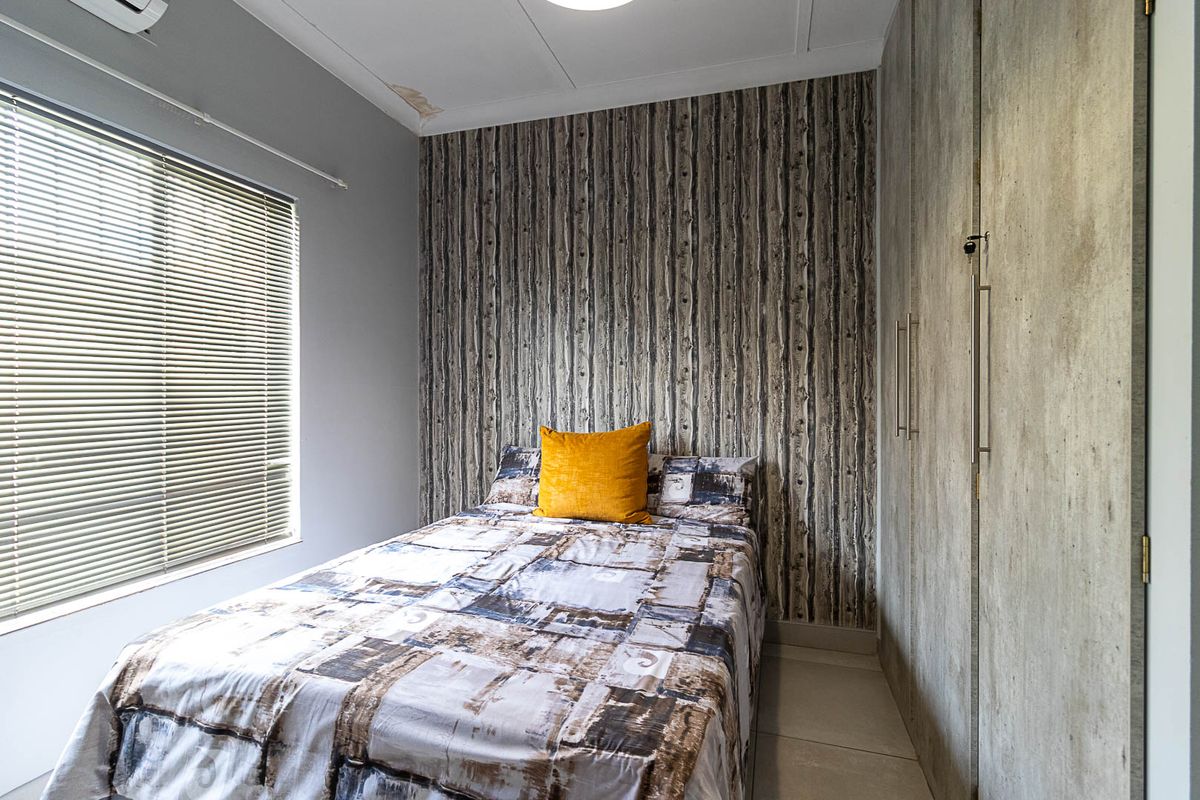 A compact bedroom featuring built-in cupboards, tiled flooring, venetian blinds, and an air conditioning unit, with visible ceiling water staining.