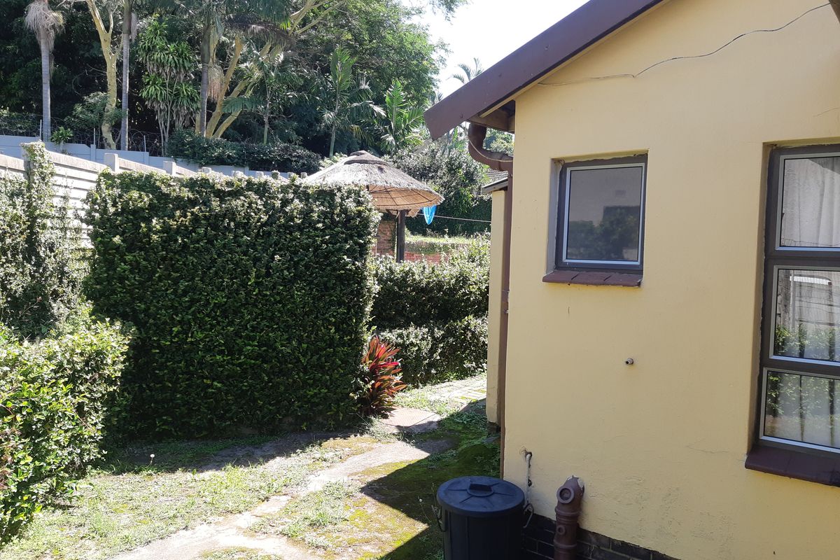 Rear exterior view of the property featuring a garden pathway, established hedges, and a thatched gazebo in the background.