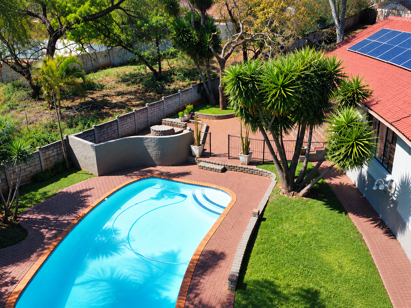 Aerial view of a backyard featuring a swimming pool, paved patio area, landscaped garden, and solar panels installed on the roof.