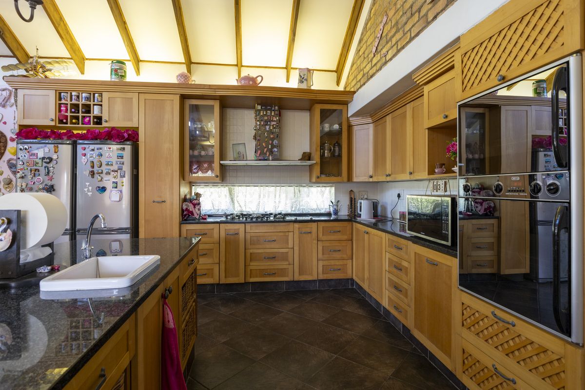 Spacious kitchen featuring warm wooden cabinetry, granite countertops, a gas hob, built-in oven, and striking exposed timber beams with a feature brick wall.
