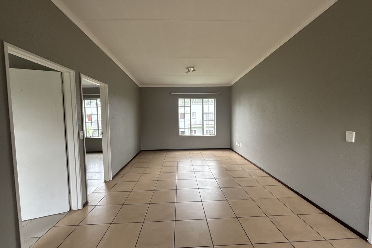 A spacious, empty living area featuring neutral grey walls, ceramic tiled flooring, and a large window providing natural light.
