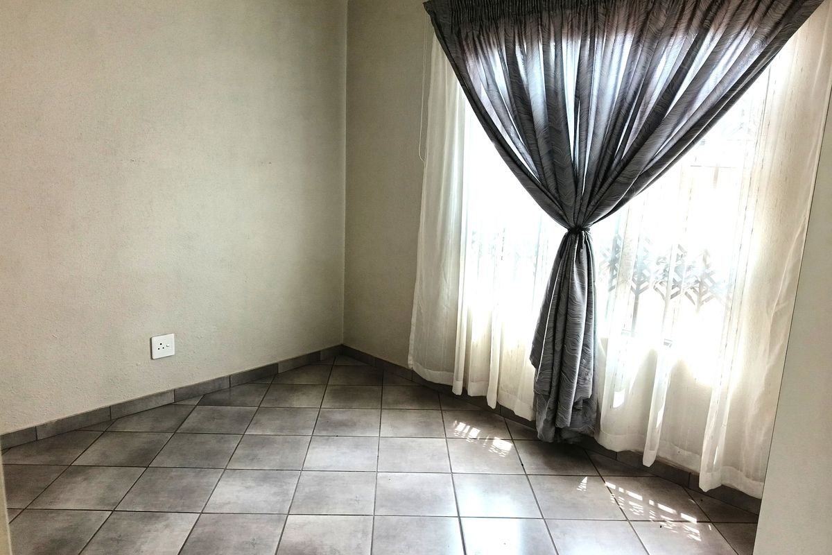 A bright, empty bedroom featuring neutral wall tones, tiled flooring, and a large window dressed with curtains.