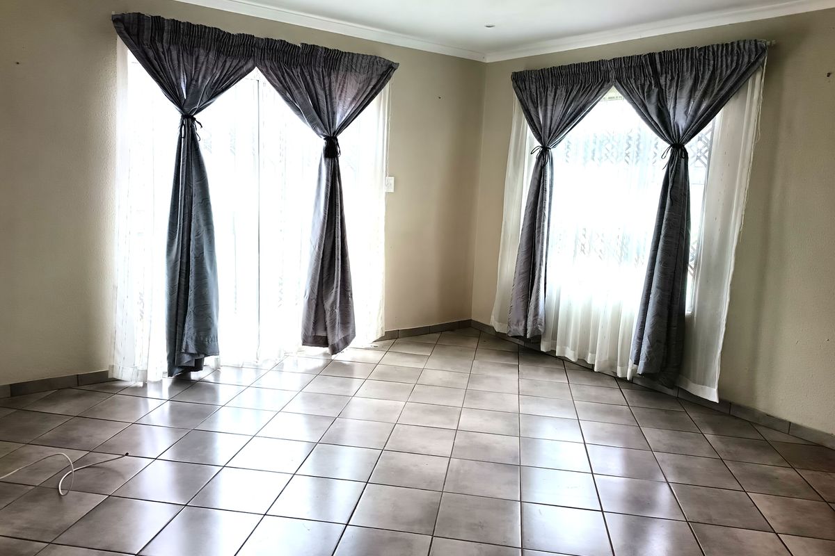 A bright, empty living room featuring neutral ceramic floor tiles, large windows with natural light, and coved ceiling cornicing.